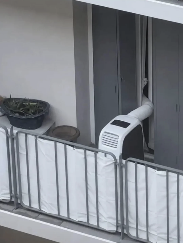Climatiseur portable blanc et noir posé sur un balcon avec sa gaine d'évacuation passant par une fenêtre ouverte dans un immeuble, à côté d'un panier contenant des plantes.