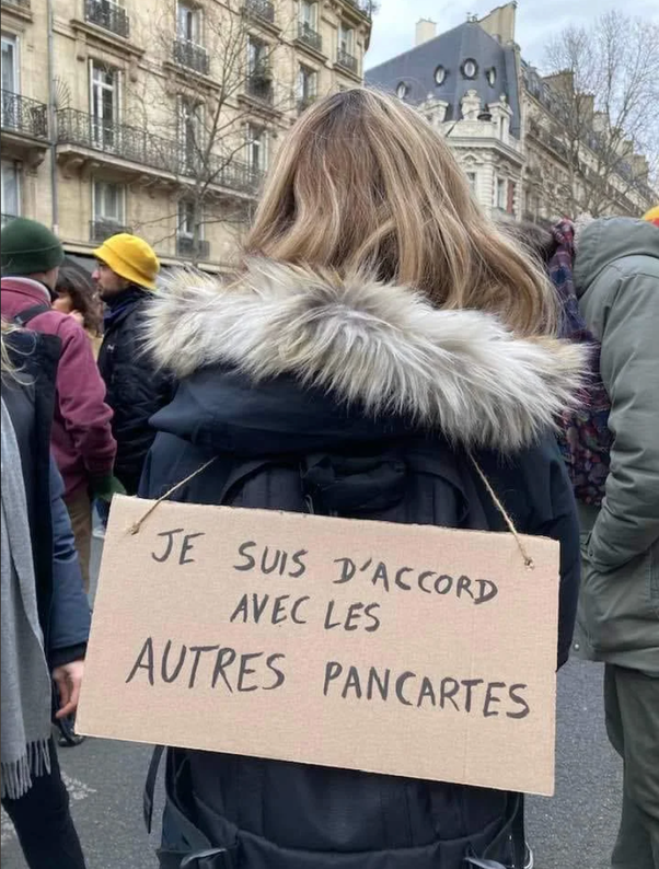 Personne vue de dos dans une rue de Paris, portant un manteau noir avec col en fourrure blanche et un sac à dos noir. Une pancarte en carton accrochée au sac affiche le texte 'JE SUIS D'ACCORD AVEC LES AUTRES PANCARTES'. Arrière-plan avec d'autres manifestants et immeubles haussmanniens.