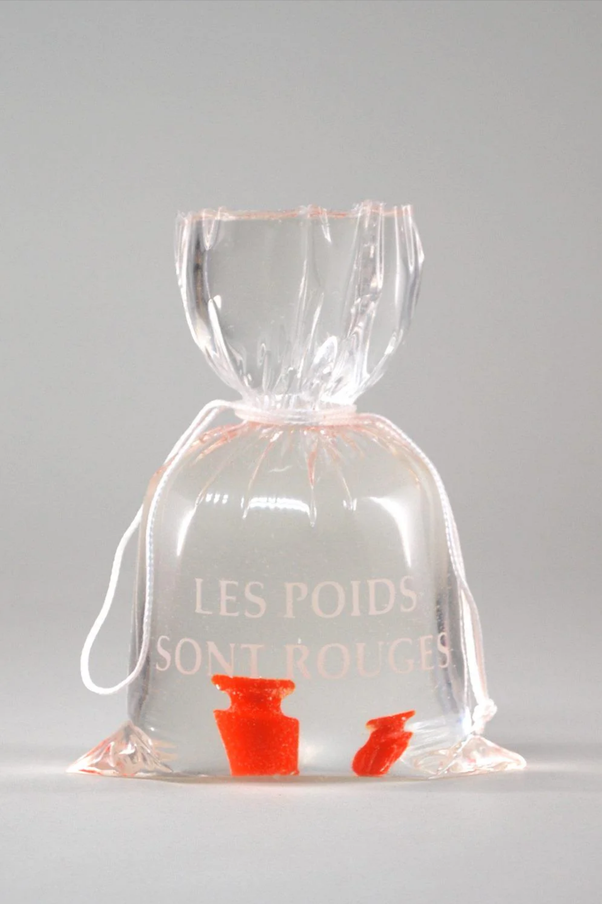 Sac en plastique transparent noué par une ficelle blanche, portant le texte blanc LES POIDS SONT ROUGES, avec deux petits objets rouges au fond.