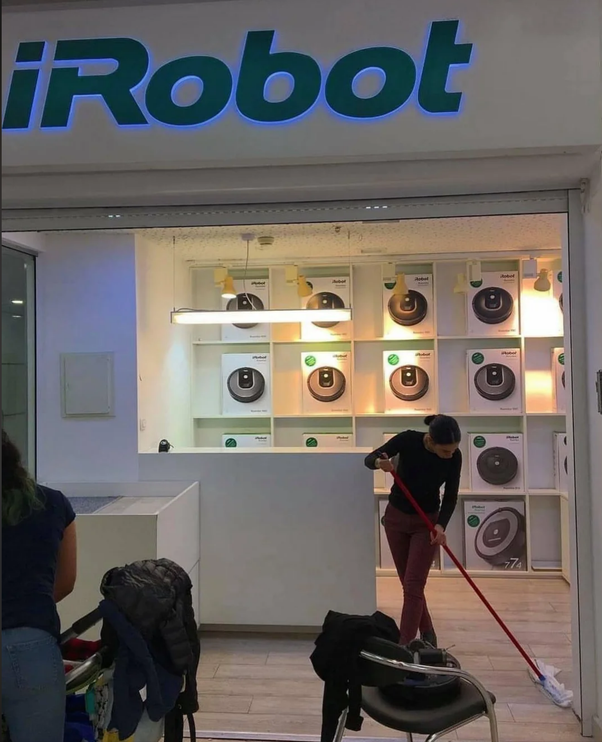 Une femme en pull noir et pantalon rose passe une serpillière rouge dans l'entrée d'un magasin iRobot avec des aspirateurs robots exposés sur des étagères et le logo iRobot en grand.
