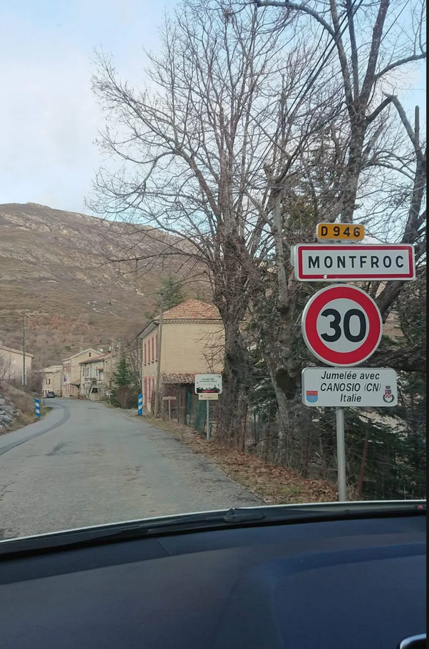 Panneau routier blanc et rouge 'MONTFROC' avec panneau de limitation de vitesse à 30 km/h et panneau 'Jumelée avec CANOSIO (CN) Italie' avec drapeaux, vu depuis l'intérieur d'une voiture sur une route de campagne bordée de maisons, arbres dénudés et collines.