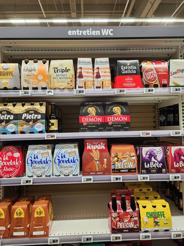 Rayons de supermarché chargés de packs de bières diverses sous le panneau 'entretien WC'.