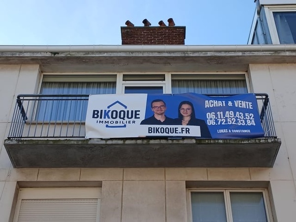 Pancarte bleue et blanche de Bikoque Immobilier avec les portraits de Lukas et Constance, logo, numéros de téléphone et texte achat vente, suspendue sur le balcon d'un immeuble beige avec des pigeons sur la cheminée.