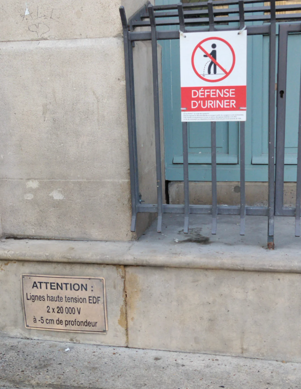 Panneau rouge et blanc avec pictogramme d'interdiction d'uriner et texte 'DÉFENSE D'URINER', au-dessus d'une plaque 'ATTENTION : Lignes haute tension EDF 2 x 20 000 V à -5 cm de profondeur' sur un mur en pierre devant une grille métallique.