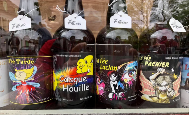 Quatre bouteilles de bière blonde artisanale alignées sur une étagère avec des étiquettes colorées représentant des fées et des personnages cartoon. Noms : Fée Tarde, Casque Houille, La Fée Lacion et La Fée Pachier. Tags 'féo' suspendus à chaque goulot.