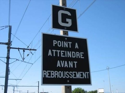 Panneau noir rectangulaire avec bord blanc et texte blanc en majuscules 'POINT A ATTEINDRE AVANT REBROUSSEMENT'. Au-dessus, petit panneau carré noir avec 'G' blanc. Monté sur poteau métallique sous ciel bleu clair avec câbles et poteaux en fond.