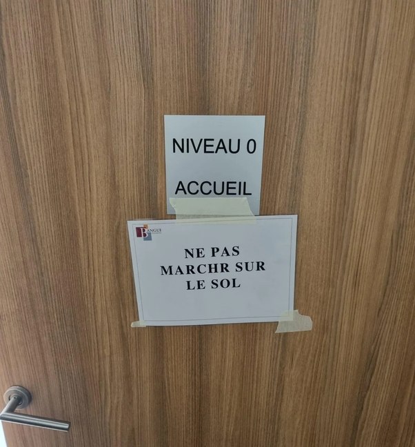 Porte en bois avec deux panneaux blancs fixés par du ruban adhésif. Haut : NIVEAU 0 ACCUEIL. Bas : NE PAS MARCHER SUR LE SOL avec un logo danger.