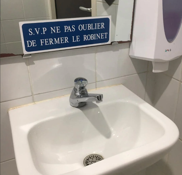 Lavabo blanc avec robinet en chrome dans une salle de bain aux murs carrelés blancs. Au-dessus, un panneau bleu porte l'inscription 'S.V.P NE PAS OUBLIER DE FERMER LE ROBINET'. Un distributeur de savon blanc est visible sur le côté droit.