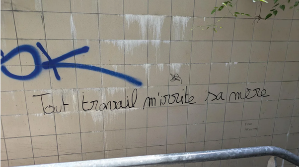 Mur de carreaux beiges avec taches blanches, symbole bleu et texte manuscrit 'Tout travail m'énerve sa mère' en noir, rambarde métallique en bas.