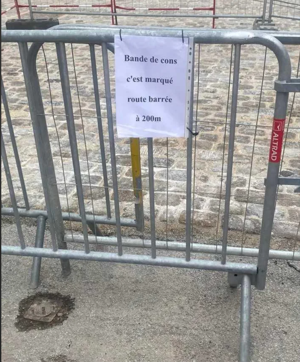 Barrière de chantier en métal avec un panneau blanc attaché affichant 'Bande de cons c'est marqué route barrée à 200m' sur un sol en pavés.