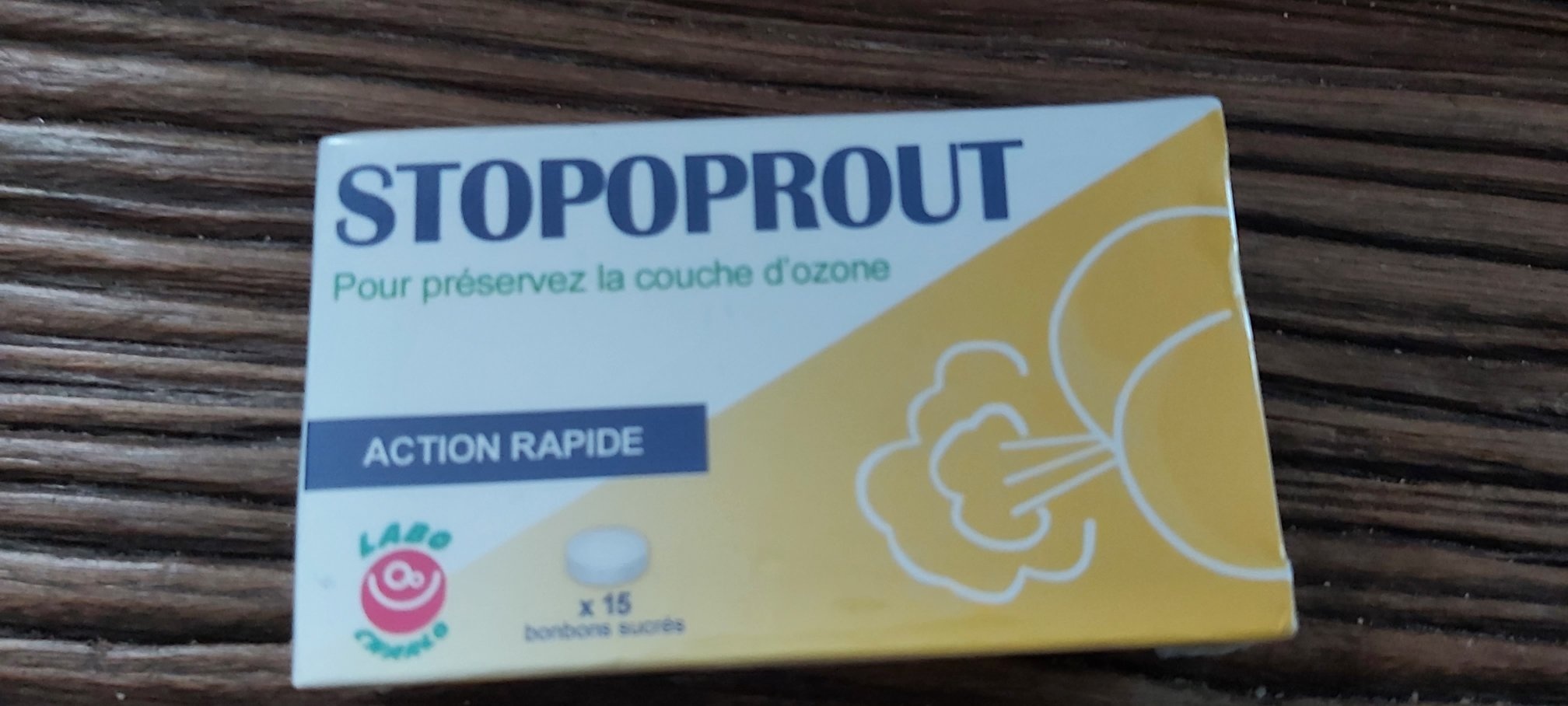 Boîte blanche et jaune de bonbons Stopoproute avec inscriptions 'STOPOPROUT', 'Pour préserver la couche d'ozone' et 'ACTION RAPIDE', logo labo et illustration, posée sur du bois.
