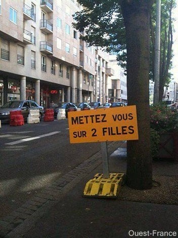 Panneau jaune rectangulaire avec le texte METTEZ VOUS SUR 2 FILLES fixé à un arbre au bord d'une rue urbaine avec immeubles et voitures en arrière-plan.