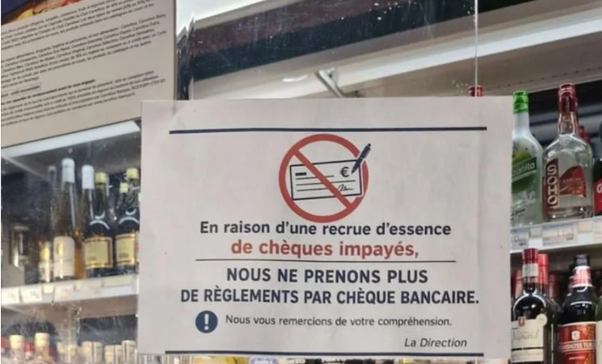 Panneau blanc dans un magasin affichant un symbole de chèque barré et le texte refusant les règlements par chèque bancaire en raison de chèques impayés, avec des bouteilles d'alcool en arrière-plan.