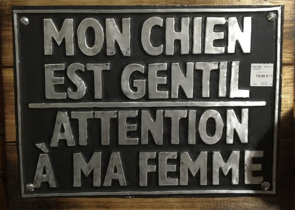 Panneau en métal noir avec texte argenté en relief : MON CHIEN EST GENTIL au-dessus d'une ligne, ATTENTION À MA FEMME en dessous. Monté sur un panneau de bois.