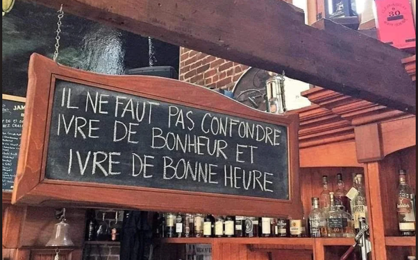 Panneau encadré en bois suspendu dans un bar avec des bouteilles d'alcool en arrière-plan. Inscription à la craie sur tableau noir : IL NE FAUT PAS CONFONDRE IVRE DE BONHEUR ET IVRE DE BONNE HEURE.