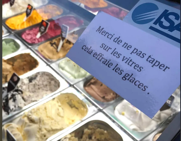 Une vitrine de glaces artisanales présentant de nombreux parfums dans des bacs métalliques rectangulaires, accompagnée d'une pancarte indiquant 'Merci de ne pas taper sur les vitres cela effraie les glaces' avec un logo bleu ISA en haut.