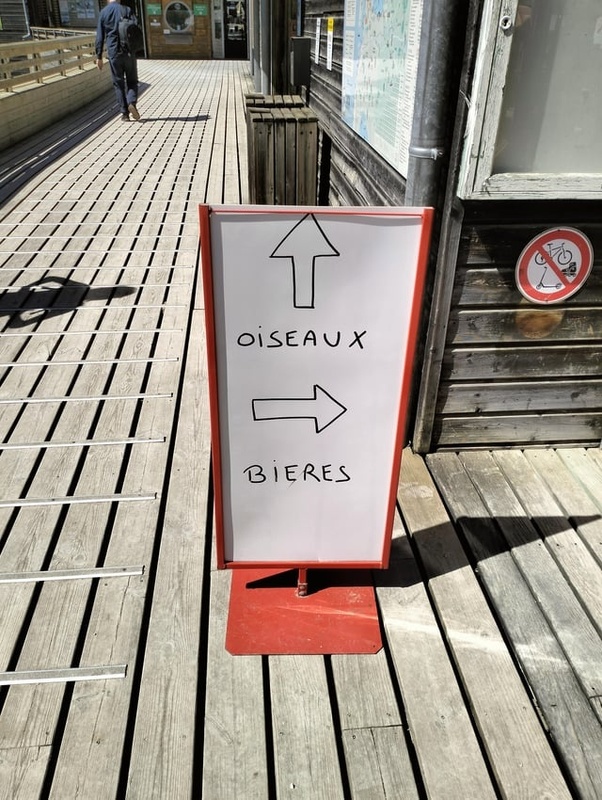 Panneau blanc avec bord rouge sur terrasse en bois, flèche vers le haut 'OISEAUX' et flèche vers la droite 'BIERES'. Panneau interdit vélos sur mur en bois et personne en arrière-plan.
