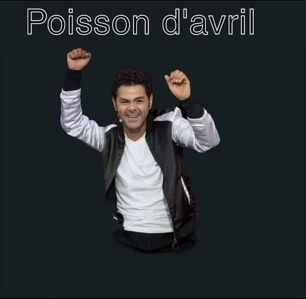 Photomontage d'un homme aux cheveux bouclés sombres, souriant, en t-shirt blanc et veste noire à manches blanches, bras levés poings serrés, avec le texte 'Poisson d'avril' en haut sur fond noir.