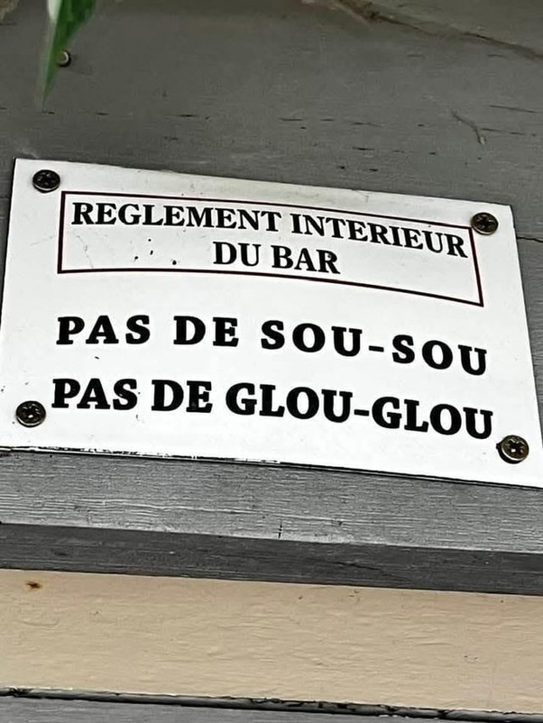 Panneau blanc rectangulaire vissé sur un mur en bois gris, texte en majuscules noires : REGLEMENT INTERIEUR DU BAR, PAS DE SOU-SOU, PAS DE GLOU-GLOU.