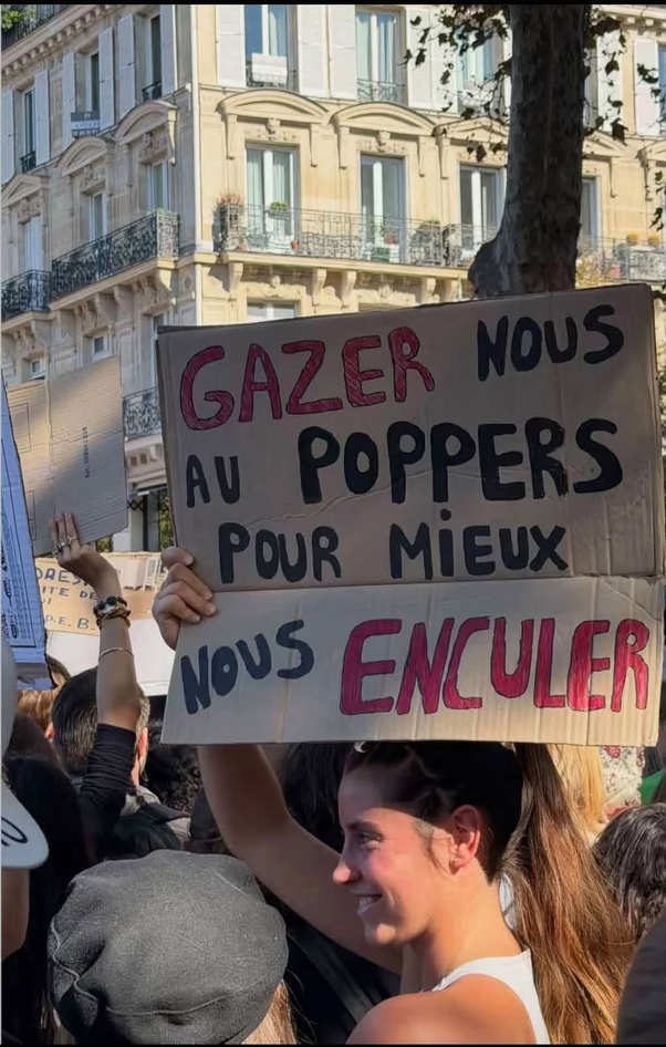 Une jeune femme à la queue de cheval tenant une pancarte en carton divisée en deux parties lors d'une manifestation à Paris. Le texte en haut est 'GAZER NOUS AU POPPERS POUR MIEUX' et en bas 'NOUS ENCULER', écrit en noir et rouge sur fond beige.