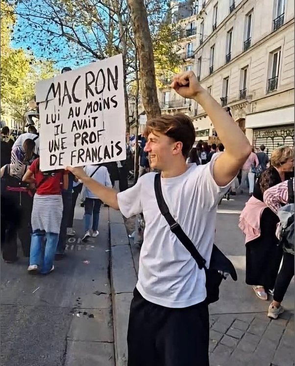 Jeune homme en t-shirt blanc tenant une pancarte avec le texte MAC RON LUI AU MOINS IL AVAIT UNE PROF DE FRANÇAIS, poing levé, dans une rue de Paris.