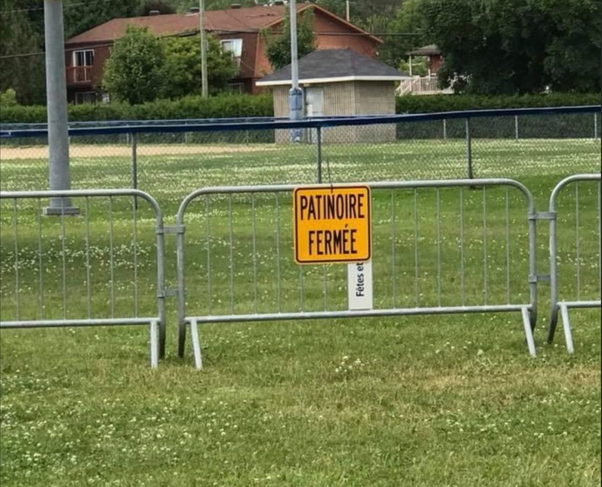 Panneau jaune indiquant PATINOIRE FERMÉE devant des barrières sur un champ d'herbe avec fleurs blanches, clôture et bâtiments en arrière-plan.